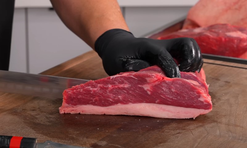 A person wearing black gloves skillfully cuts a piece of meat on a cutting board