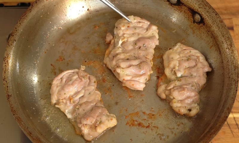 Three pieces of chicken are sizzling in a frying pan on a stovetop
