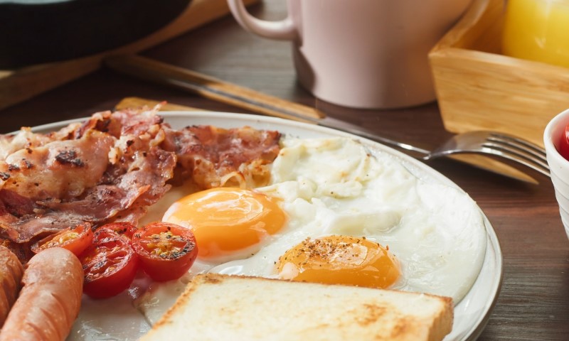 A breakfast plate with crispy bacon, two sunny-side-up eggs, grilled tomatoes, toast, and a sausage